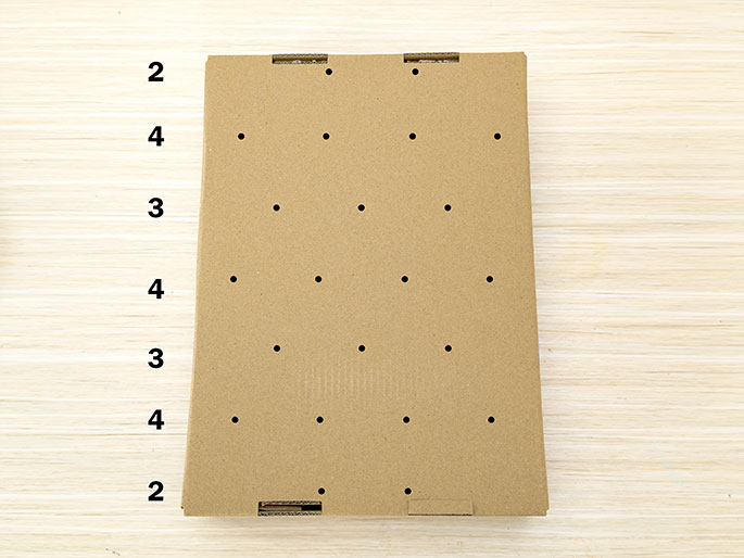 A rectangular piece of flat cardboard rests on a light wooden surface. The cardboard has a grid of small, evenly spaced holes punched in it. The numbers 2, 4, and 3 are written repeatedly along the left edge of the cardboard, likely indicating measurements or a pattern for a craft or game.