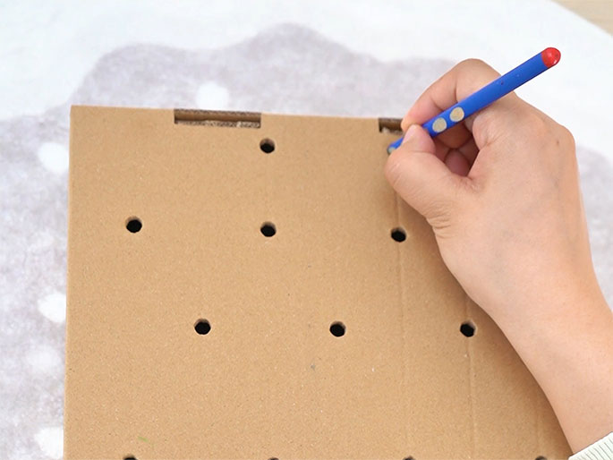 A hand holding a blue crayon colors one of several pre-punched holes in a piece of light brown cardboard. The cardboard sits on a white textured surface. The image suggests a craft activity in progress.