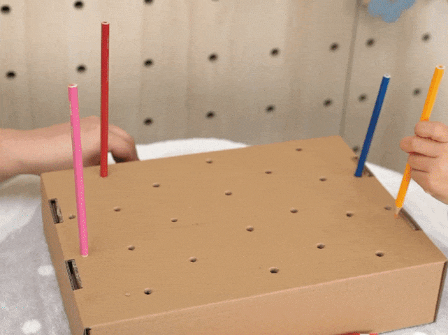 Children's hands are inserting colored pencils—pink, red, blue, and yellow—into the pre-punched holes of a piece of light brown cardboard. The cardboard rests on a white textured surface. This image depicts a playful and creative activity, likely a craft or game involving color and coordination.