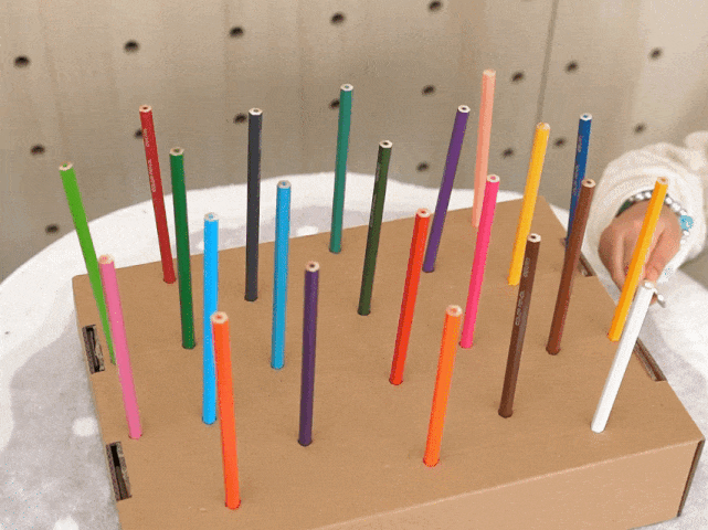 A piece of cardboard with pre-punched holes is filled with an array of upright colored pencils. The pencils vary in color, including shades of green, pink, red, blue, orange, purple, gray, brown, and white. A child's hand is visible in the bottom right corner, placing a yellow pencil into one of the holes. The cardboard sits on a white textured surface. The image captures a colorful and engaging activity, likely a craft or game designed for children.