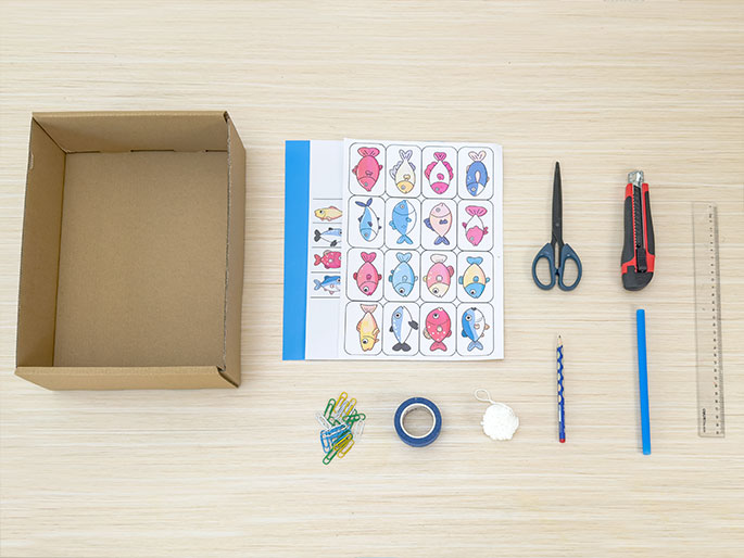 An overhead flatlay image displays various craft supplies arranged on a light wood surface. These include an open cardboard box, a sheet of paper with printed fish images, scissors, a utility knife, a ruler, paper clips, blue tape, a small ball of white yarn or string, a pencil, and a blue plastic straw or stick. The arrangement suggests preparation for a children's craft project, possibly a fishing game.