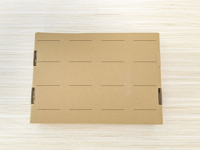 A rectangular piece of flat cardboard lies on a light wooden surface. Horizontal lines are drawn across the cardboard, dividing it into equal rectangular sections. The edges of the cardboard show slight creases or folds, suggesting it may have been part of a box. The image evokes a sense of preparation for a craft or organizational project.