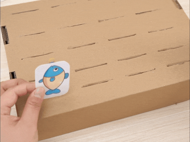 A hand holds a small, square piece of paper with a drawing of a simple, cartoon fish on it. The paper appears to have a small metal brad or fastener at the top. The hand is positioning the fish over a larger piece of cardboard with several thin, parallel slits cut into it. The background is a light wood surface. The image suggests a fishing game or a child's craft activity.