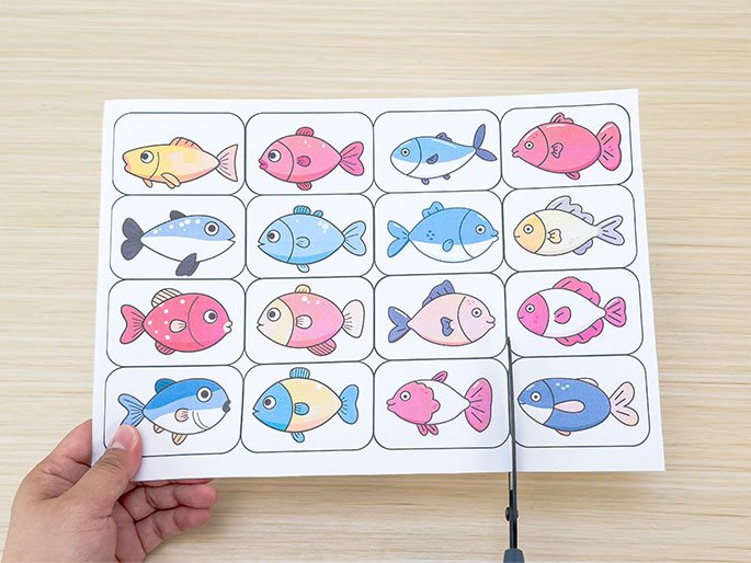 A hand holds a sheet of paper with sixteen printed images of colorful cartoon fish, arranged in a grid. Scissors are positioned to cut the paper, separating the individual fish images. The background is a light wooden surface. The image suggests the preparation of game pieces or craft elements for a children's activity.