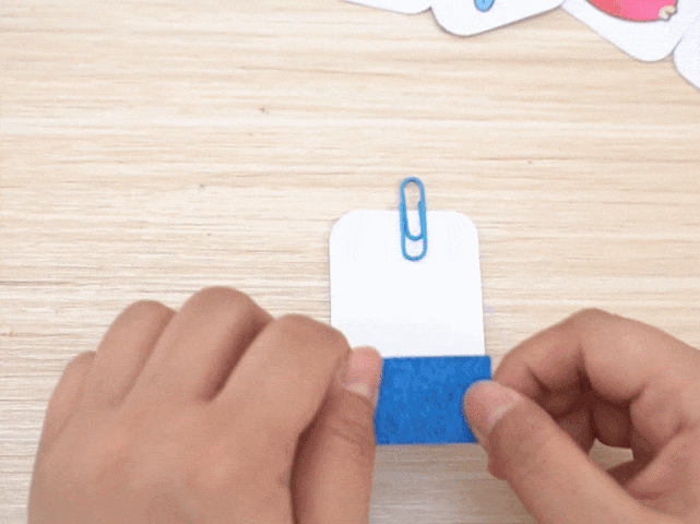Two hands are shown attaching a strip of blue textured material to the bottom edge of a small white card. A light blue paperclip is clipped to the top of the card. The background is a light wood surface. The image suggests someone creating a handmade element, possibly for a game or craft project.