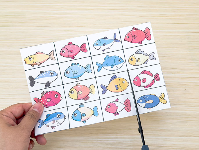 A hand holds a piece of paper containing sixteen colorful cartoon fish illustrations arranged in a grid. Scissors are cutting the paper, separating the fish images. The fish are various colors, including pink, yellow, blue, and orange. The background is a light wooden surface. The scene suggests the preparation of pieces for a children's game or a crafting activity.