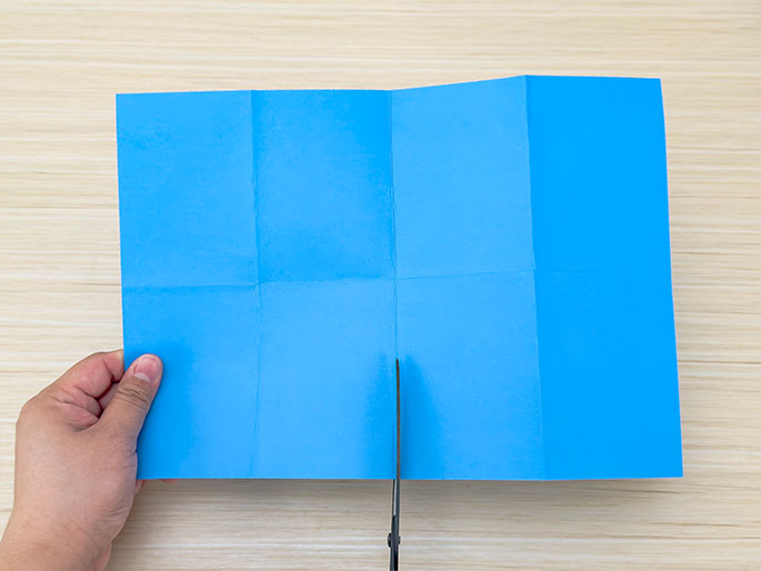 A hand holds a piece of bright blue paper folded into fourths. Scissors are positioned to cut the paper along one of the central folds. The paper rests on a light wood surface. The image suggests a simple paper-cutting activity or the preparation of materials for a craft project.