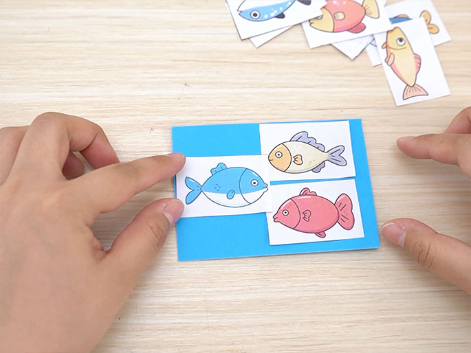 Two hands are placing small, printed images of colorful cartoon fish onto a larger piece of light blue cardstock. A blue and white fish is being positioned next to pre-placed images of a yellow and white fish and a pink fish. Additional fish images are scattered near the top of the frame. The background is a light wooden surface. The image suggests a matching game or a crafting activity involving fish.