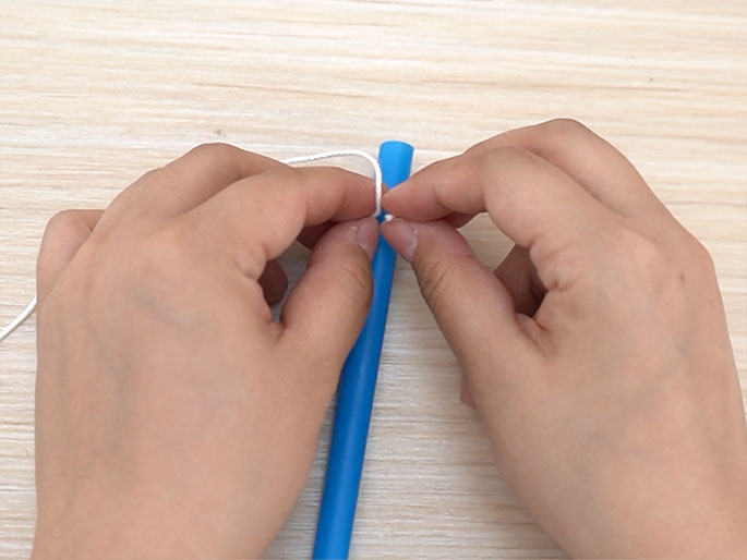 Two hands are tying a piece of white string or yarn around a blue plastic drinking straw. The hands are in the process of tightening the knot. The background is a light wood surface. The image suggests a crafting activity or the creation of a homemade toy or tool.