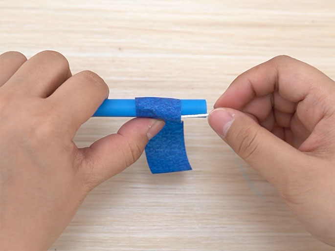 Two hands are shown working with a blue plastic drinking straw. Blue painter's tape is being wrapped around one end of the straw. A thin, white string or wire is being threaded through the end of the straw. The background is a light wood surface. The image suggests a crafting or DIY project, possibly creating a toy or tool.