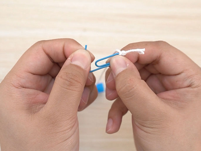 Two hands hold a small, light blue paperclip. A piece of white string or yarn is being tied to the paperclip. The background is a light wood surface. The image suggests the beginning stages of a craft project or the creation of a simple tool or toy.
