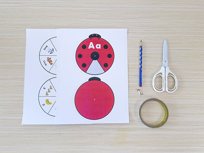 The image shows the materials for a ladybug-themed children's learning activity arranged on a light wood-grain table. Two printed sheets display ladybug parts and spinner wheels. One spinner features images of a bird, fish, and ball, each labeled with the corresponding word. The other spinner displays numbers and matching quantities of bananas and pizza slices. A pencil, scissors, a brass fastener, and a roll of clear tape are placed beside the printed sheets. These materials suggest a craft or game involving assembling the ladybug and using the spinners, likely for literacy and numeracy development.