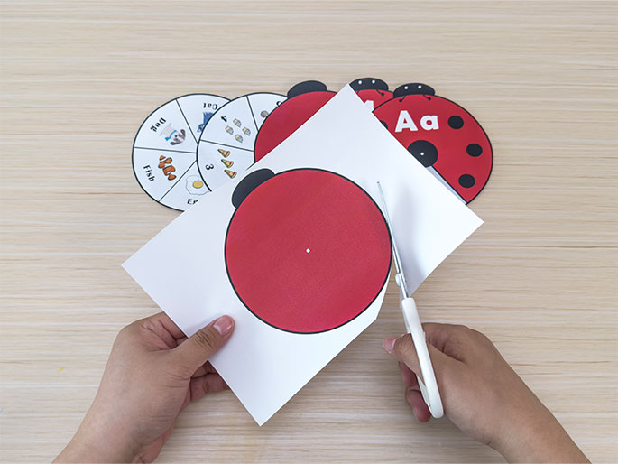 Two hands are shown cutting out a red circle from a piece of paper using white-handled scissors. The red circle is part of a ladybug craft, as other ladybug components and learning spinners are visible on the light wood-grain table. The spinners are divided into segments showing pictures of a dog, bird, fish, egg, and quantities of pizza slices and bananas, along with corresponding words and numbers. This image captures the preparation stage of a ladybug-themed children's activity, likely focused on developing early literacy and numeracy skills.