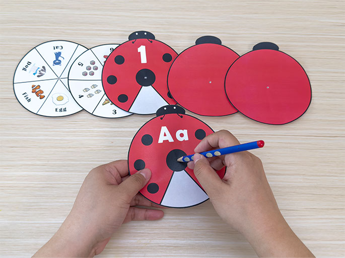 Two hands are shown assembling a ladybug learning aid on a light wood-grain table. A pencil is being used to poke a hole in the center of a red and black ladybug cutout with the letters Aa printed on it. Other ladybug parts and learning spinners are visible on the table. The spinners are divided into segments, one showing pictures of a dog, bird, fish, egg, and quantities of pizza slices and ice cream cones, along with corresponding words and numbers. This image captures the construction phase of a children's educational activity focused on basic literacy and numeracy skills.