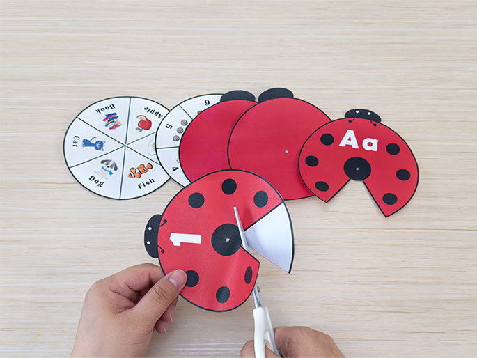 Two hands are using scissors to cut a ladybug-shaped learning aid on a light wood-grain table. The ladybug is red with black spots and has the number 1 printed on it. Other ladybug parts and a learning spinner are visible on the table. The spinner is divided into segments showing pictures of a cat, dog, fish, book, apple, along with corresponding words. This image captures the process of creating a children's educational game focused on basic literacy and numeracy.