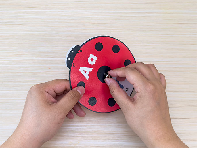Two hands assemble a ladybug-shaped learning tool on a light wood-grain table. One hand holds the red and black ladybug cutout with the letters Aa printed on it, while the other hand fastens a metal brad through the center. The edge of a small picture card with the word cat is visible beneath the hand inserting the brad. This close-up image shows the final steps of creating an educational aid for children, likely designed as a spinner or matching game.