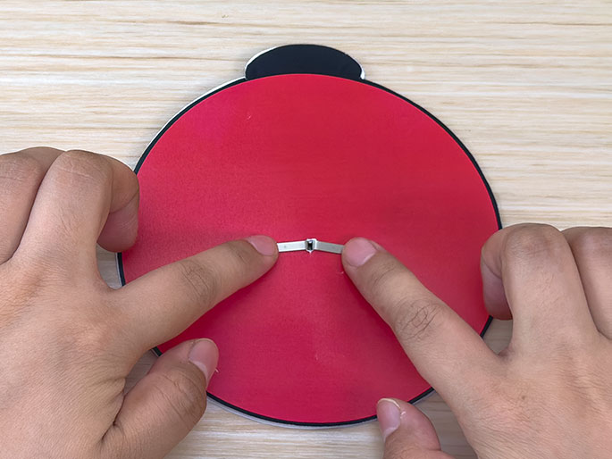 Two hands manipulate a brad fastener on the back of a red circular piece of material, likely part of a ladybug craft project. The brad's prongs are being folded down against the red surface, which has a black section at the top suggesting the ladybug's head. The hands and the red circle are positioned on a light wood-grain surface. This close-up image focuses on the assembly process of securing the brad, a common step in children's crafts.