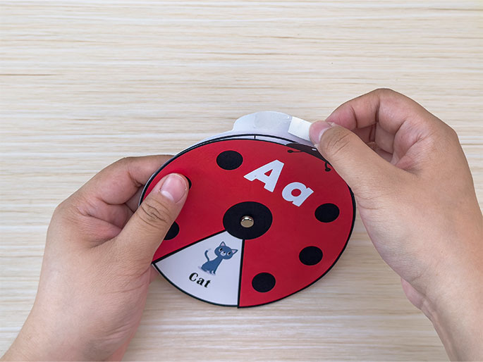 Two hands are shown assembling a ladybug-shaped learning spinner on a light wood-grain table. One hand holds the spinner, which features the letters Aa, a picture of a cat, and the word Cat. The other hand peels back a piece of double-sided tape from the spinner's edge. This image illustrates the process of creating an interactive educational tool for children.