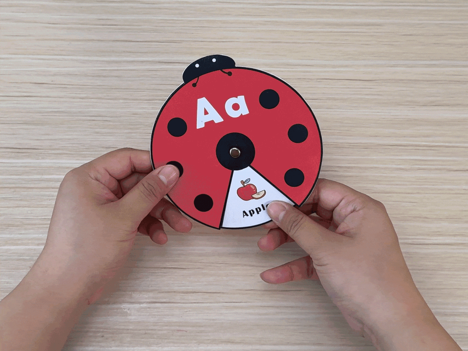 Two hands hold a circular ladybug-themed learning toy. The toy features the letter Aa and rotates to reveal corresponding words and pictures, such as Apple.