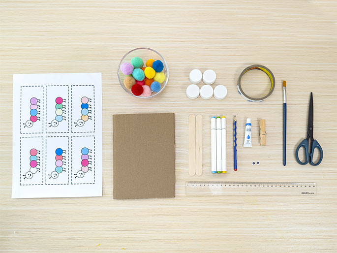 An assortment of craft supplies is laid out on a light wood table. These include: a sheet of paper with printable caterpillar segments, a bowl of colorful pom-poms, several white bottle caps, a roll of clear tape, a paintbrush, scissors, large craft sticks, white markers, a pencil, a tube of blue paint, a clothespin, googly eyes, and a clear plastic ruler. The arrangement suggests preparation for a children's craft project involving caterpillars.