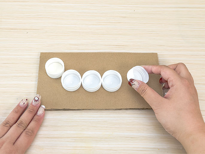 Two hands are arranging white plastic bottle caps on a piece of light brown cardboard. One hand is placing a cap onto the cardboard, while the other hand rests near the edge. Four caps are already lined up on the cardboard. The background is a light wood surface. The image suggests a crafting activity or the beginning stages of a DIY project.