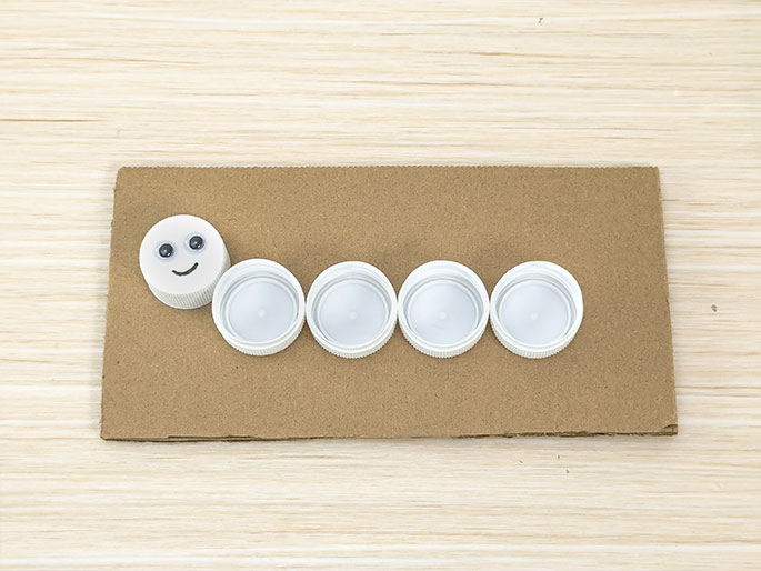 Five white plastic bottle caps are arranged on a piece of light brown cardboard to resemble a caterpillar. The leftmost cap has googly eyes and a drawn smile, representing the caterpillar's head. The other four caps form the body. The image is set against a light wood background. This depicts a simple craft project made from recycled materials.