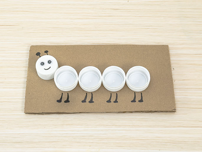 A simple caterpillar craft is depicted on a light wood background. Five white plastic bottle caps are glued to a piece of light brown cardboard. The leftmost cap, the head, has googly eyes, a drawn smile, and small antennae. Drawn black legs extend beneath each bottle cap segment. This image showcases a children's craft activity using recycled materials.