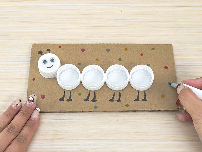 A hand with a marker is adding colorful dots to a piece of light brown cardboard. On the cardboard is a caterpillar craft made from five white plastic bottle caps. The leftmost cap, the head, has googly eyes, a drawn smile, and antennae. Black legs are drawn beneath each bottle cap. The image shows the process of decorating the caterpillar craft against a light wood background.