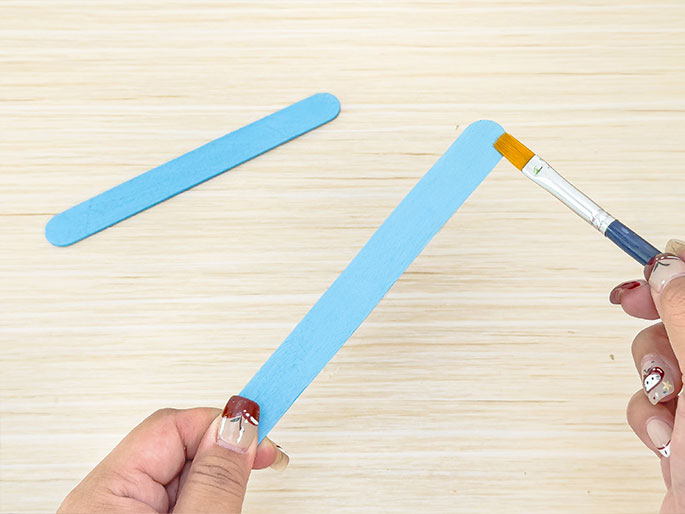 Two hands are painting light blue craft sticks a brighter shade of blue. One stick is fully painted and lies on a light wood surface, while the other is being painted with a small brush. The image focuses on the process of painting the craft sticks.