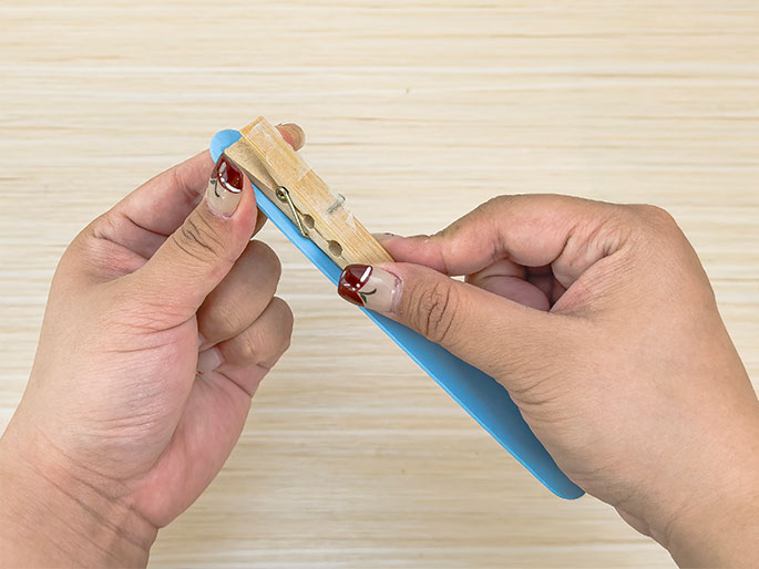 Two hands are clamping a small wooden clothespin onto a light blue craft stick. The clothespin is positioned near the rounded end of the stick. The background is a light wood surface. This image suggests a step in a crafting process.