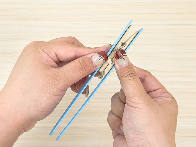 Two hands hold two light blue craft sticks with a small wooden clothespin clamped between them. The clothespin is positioned near the rounded ends of the sticks. The background is a light wood surface. This image likely demonstrates a step in assembling a craft project.