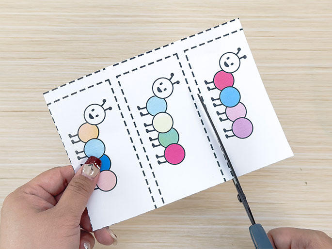 A pair of scissors is cutting apart a piece of paper with three colorful caterpillar drawings on it. Each caterpillar is made of different colored circles and has a simple face and antennae. The paper has dashed lines indicating where to cut, separating the caterpillars. The background is a light wood surface. This image likely depicts a step in preparing a children's craft or learning activity.