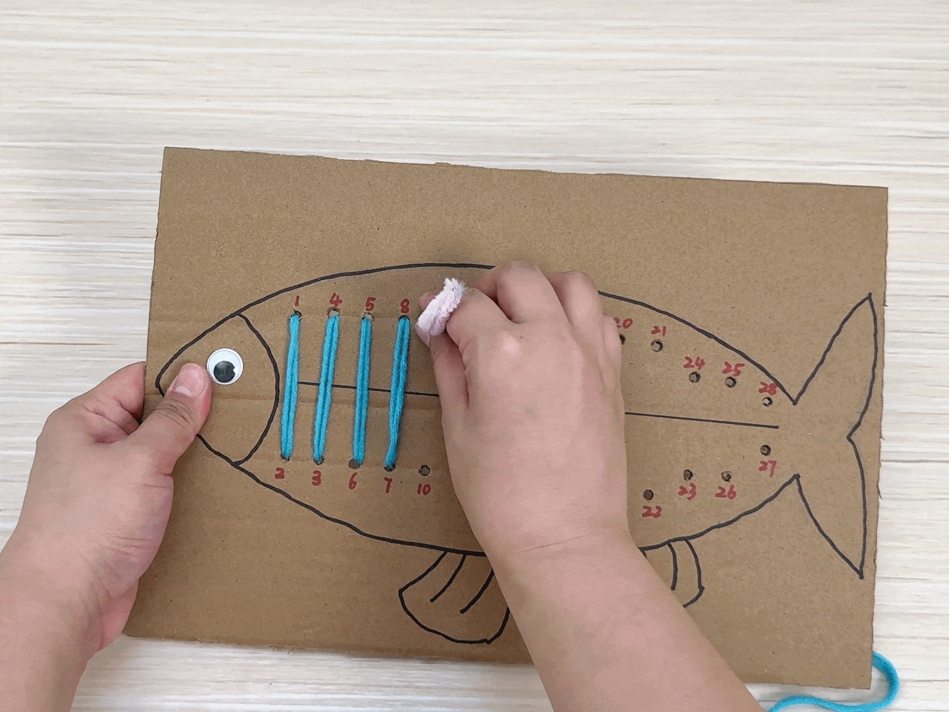 Hands demonstrate a sewing activity using a cardboard cutout of a fish. Numbered holes are punched along the fish's outline, and yarn is threaded through the holes, creating a stitching effect.