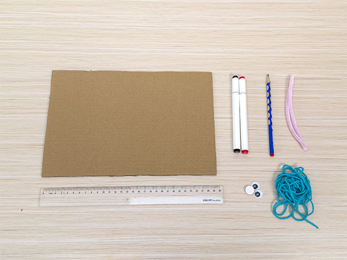 An assortment of craft supplies lies arranged on a light wood-grain table, suggesting preparation for a children's art project. The items include a rectangular piece of brown cardboard, two white dry-erase markers (one with a black cap, the other with a red cap), a blue pencil, a light pink pipe cleaner bent into a curve, a clear plastic ruler, two small white googly eyes with black pupils, and a tangle of teal yarn or string. These materials likely will be used to create a character or decorative item.