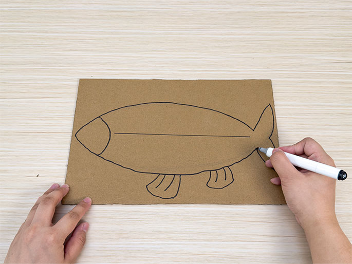 Two hands are engaged in drawing a fish on a piece of brown cardboard placed on a light wood-grain table. One hand holds the cardboard steady, while the other hand uses a white dry-erase marker to complete the outline of the fish's tail. The fish drawing features a simple, child-like style, with a large eye, a horizontal line dividing the body, small fins along the bottom, and a triangular tail. This image likely depicts a stage in a children's craft activity.