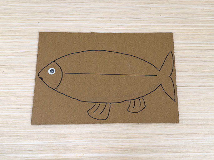 A simple fish drawing, made with black marker, is shown on a piece of brown cardboard resting on a light wood-grain surface. A small, white googly eye with a black pupil has been affixed to the fish's head. The fish is drawn in a child-like style, with a horizontal line across its body, small fins at the bottom, and a triangular tail. This image likely depicts a stage in a children's craft project where a fish is being created from cardboard.