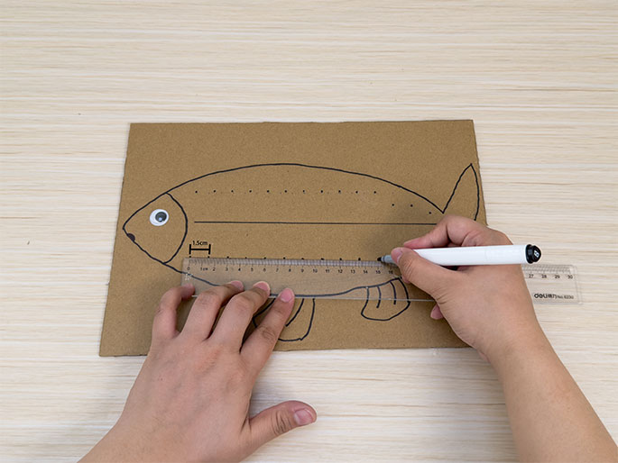 Two hands work on a fish-shaped piece of brown cardboard on a light wood-grain table. A row of dots has been drawn along the top and bottom edges of the fish outline. One hand holds a clear plastic ruler against the lower edge of the fish, while the other hand uses a white dry-erase marker to draw a straight line along the ruler. The ruler shows a measurement of approximately 1.5cm marked on the cardboard. A googly eye is already attached to the fish's head. This image seems to capture a step in a craft project, likely involving cutting or folding along the marked lines.