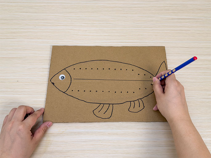A partially completed craft project featuring a fish drawn on brown cardboard sits on a light wood-grain table. The fish outline, drawn in black marker, has dotted lines along its top and bottom edges, and a solid line dividing its body horizontally. A googly eye is attached to the fish's head. One hand holds the cardboard, while the other hand uses a blue pencil to connect the dots along the fish's outline, likely in preparation for cutting or folding.