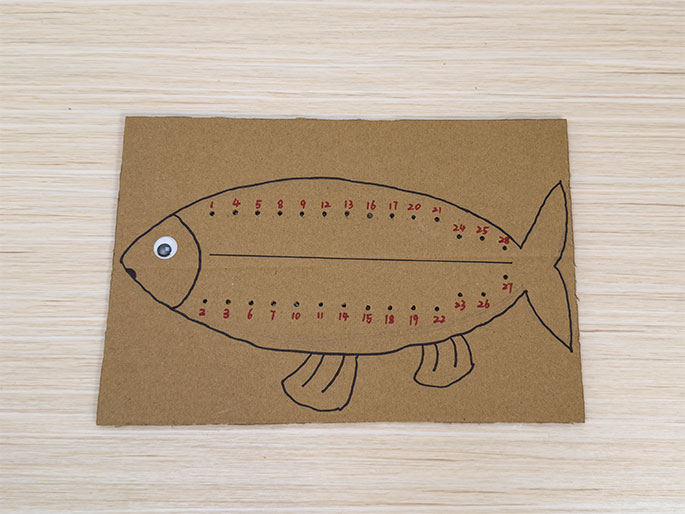 A fish shape drawn on brown cardboard is displayed on a light wood-grain table. The fish outline and fins are drawn in black marker. A googly eye is stuck to the fish's head. Small holes have been punched along the top and bottom edges of the fish's body, and red numbers are written next to each hole, sequentially from 1 to 27 along the top and 2 to 26 along the bottom. A horizontal line divides the fish's body. This image likely represents a child's sewing or lacing activity where yarn or string will be threaded through the numbered holes.