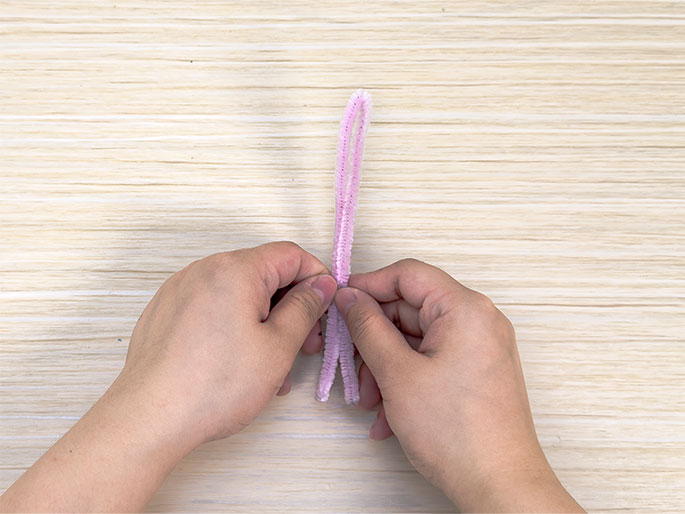 Two hands manipulate a light purple pipe cleaner against a light wood-grain background. The pipe cleaner is folded in half, and the hands are pinching the folded end together, likely as the first step in creating a shape or design with the pipe cleaner.