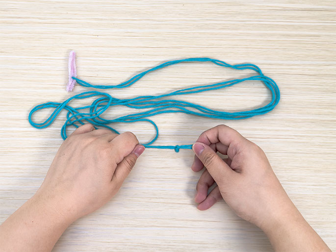Two hands hold a length of bright teal yarn against a light wood-grain background. A simple overhand knot has been tied in the yarn. A small, folded piece of light purple pipe cleaner is visible at the top left corner, suggesting it might be used later in the project. The image appears to depict a stage in a crafting activity.