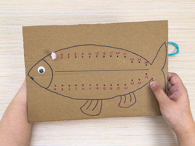 Two hands hold a piece of brown cardboard cut into the shape of a fish against a light wood-grain background. The fish has a googly eye, black outlined fins, and numbered holes punched along its top and bottom edges. A piece of light pink pipe cleaner is looped through the first few holes at the top edge. A length of teal yarn is visible at the upper right, likely to be used for lacing through the remaining holes. This image depicts a sewing or lacing activity for children.