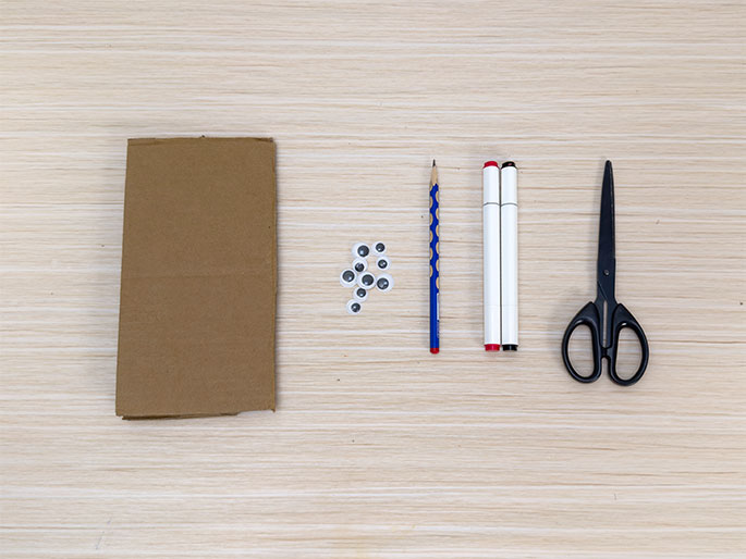 Craft supplies are arranged on a light wood-grain table. From left to right, there's a piece of brown cardboard, a small pile of googly eyes, a blue pencil, two white marker pens (one with a black cap and one with a red cap), and a pair of black scissors. These materials appear to be gathered for a children's craft project.
