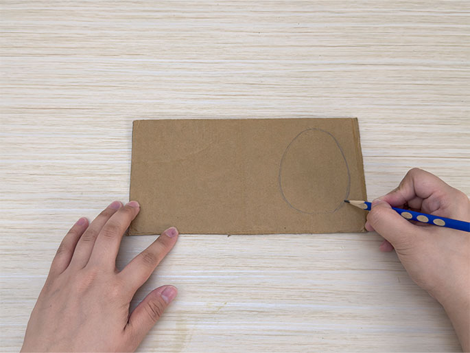 On a light wood-grain surface, two hands position a rectangular piece of brown cardboard. A blue pencil, held in the right hand, is drawing an oval shape onto the cardboard. This appears to be the beginning stage of a craft project.