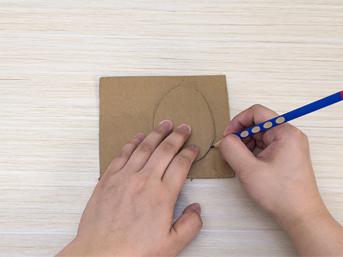 Two hands are shown on a light wood-grained table. The left hand holds down a piece of brown cardboard while the right hand uses a blue pencil to carefully draw an oval shape on the cardboard. This scene suggests the initial steps of a crafting activity.