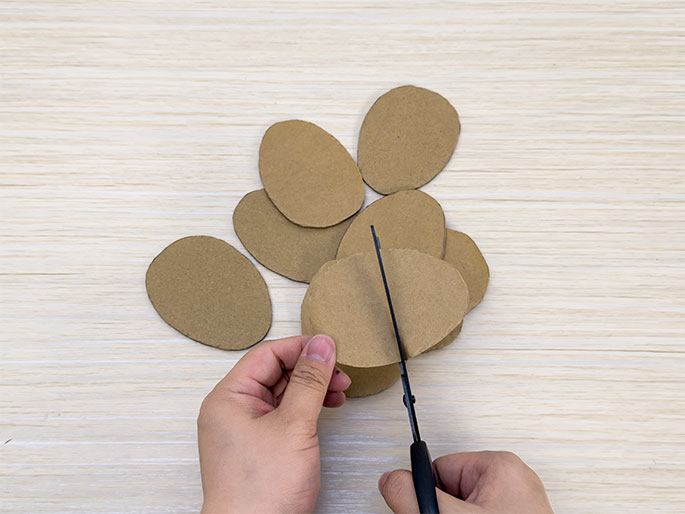 Several oval-shaped pieces of brown cardboard are scattered on a light wood-grain table. Two hands are in the frame, with one holding a piece of cardboard and the other using scissors to cut it. The action suggests the cardboard ovals are being prepared for a craft project.