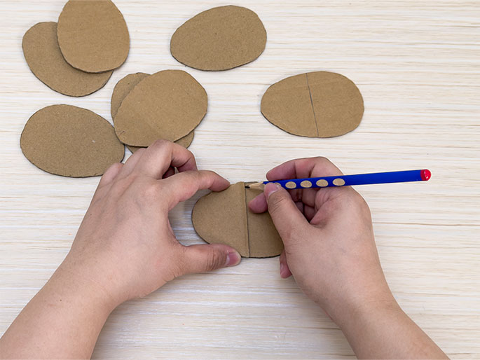On a light wood-grain table, several oval pieces of brown cardboard are visible. Two hands manipulate one of the ovals, which has a line drawn down its center. The right hand uses a blue pencil to continue drawing on the cardboard. Other similar cardboard ovals are scattered nearby. This scene suggests a craft project in progress.