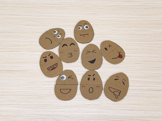 Nine oval-shaped pieces of cardboard, resembling potato shapes, are arranged on a light wood-grain table. Each cardboard potato has a different facial expression drawn on it, including happy, sad, angry, winking, surprised, and silly faces. Some have googly eyes attached. These appear to be part of a children's game or craft activity.
