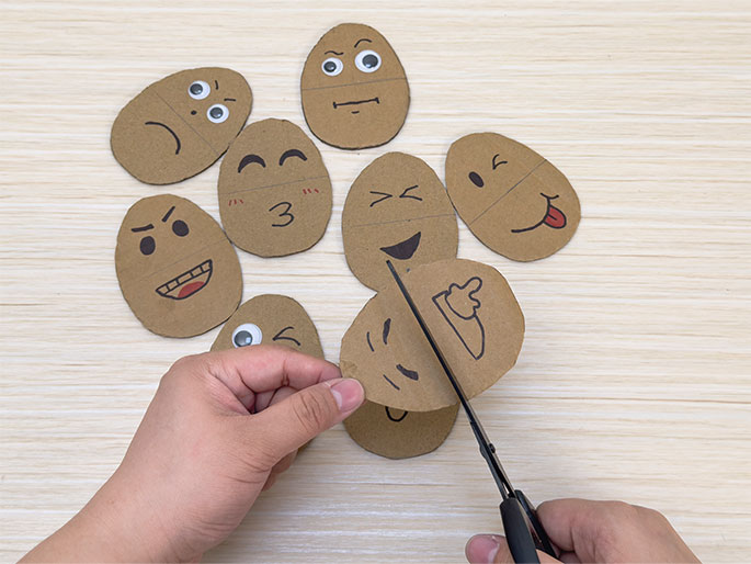 On a light wood-grain table are several oval-shaped pieces of cardboard decorated with various facial expressions, similar to emojis. Two hands are visible; one holds a cardboard oval while the other uses scissors to cut it. The oval being cut has a partial face with a winking eye and a pointing hand drawn on it. This scene is likely part of a craft activity involving creating mix-and-match faces.