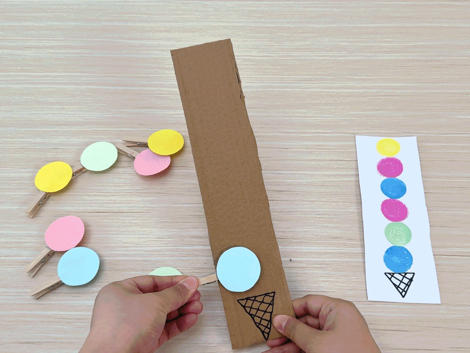 Hands clip colorful paper circles onto clothespins attached to a cardboard strip shaped like an ice cream cone. A separate piece of paper shows the intended color pattern for the ice cream scoops. This demonstrates a color-matching or patterning activity.